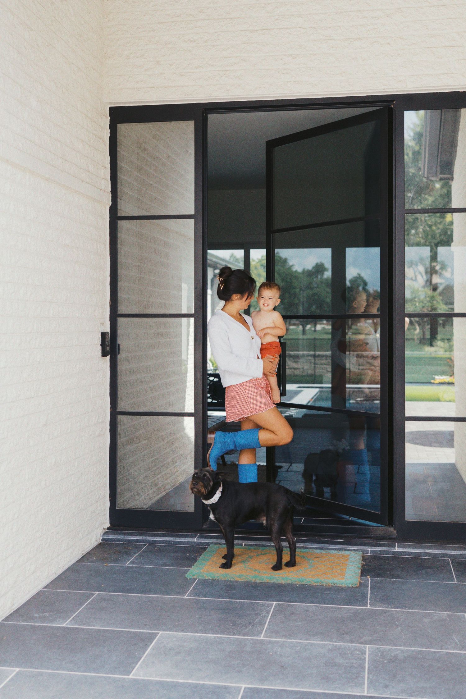 Woman holding baby stands at modern black-framed glass front door with black dog on gray slate porch.