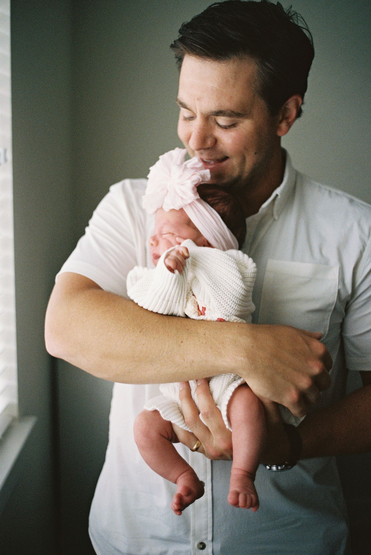 A proud father tenderly holds his newborn baby wrapped in white, smiling softly near a window.