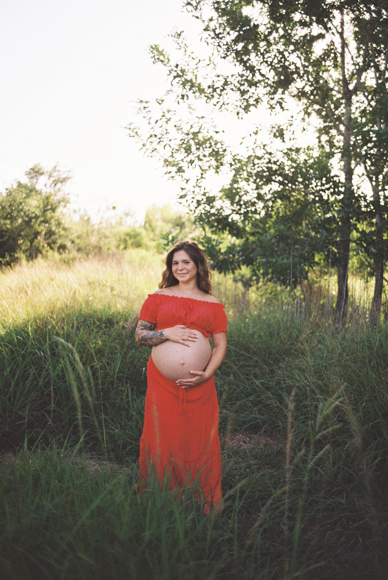 Pregnant woman in red off-shoulder dress cradling baby bump outdoors in lush green field during golden hour.