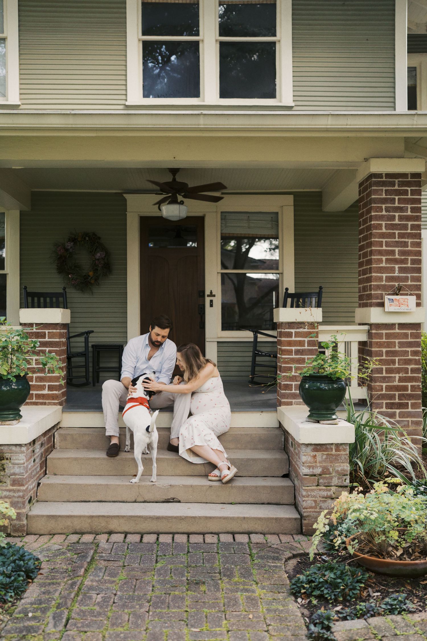 Couple sitting on front porch steps with white dog at charming brick home with ceiling fan and wreath.