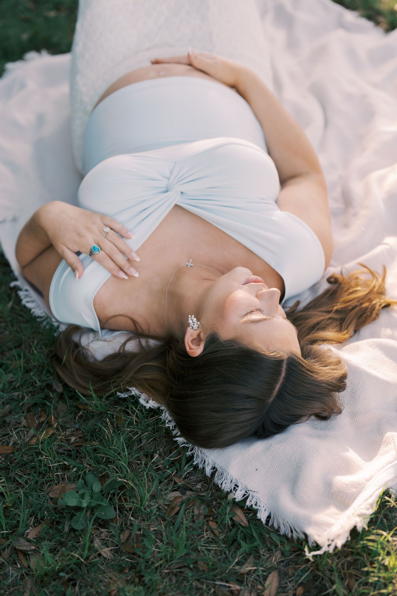 Pregnant woman in white crop top lying on blanket outdoors, cradling baby bump with hands adorned with turquoise rings.