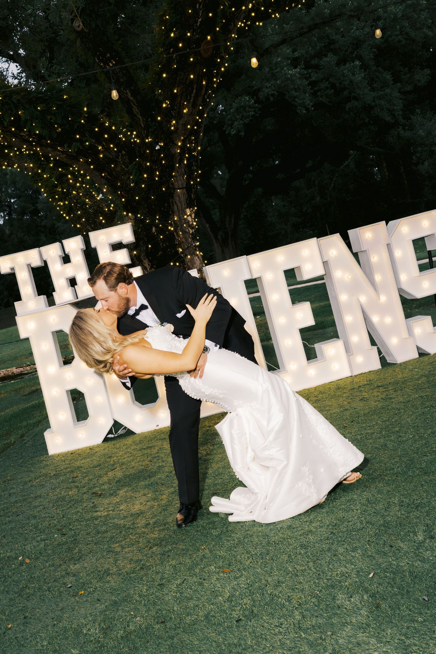 Bride and groom share a romantic dip kiss in front of illuminated marquee letter sign at outdoor wedding reception.