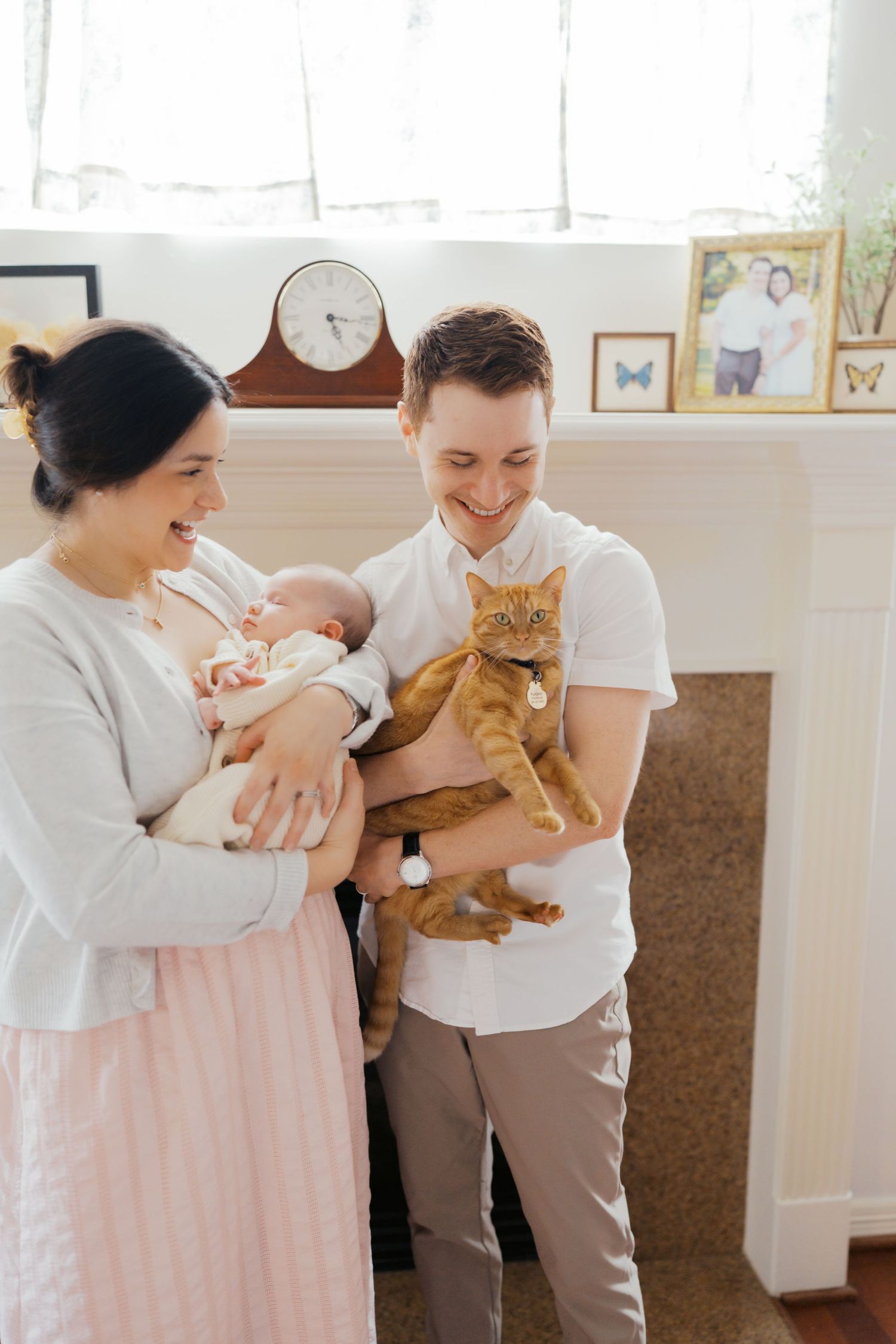 A smiling couple holds a newborn baby and an orange tabby cat in a cozy living room with a fireplace.