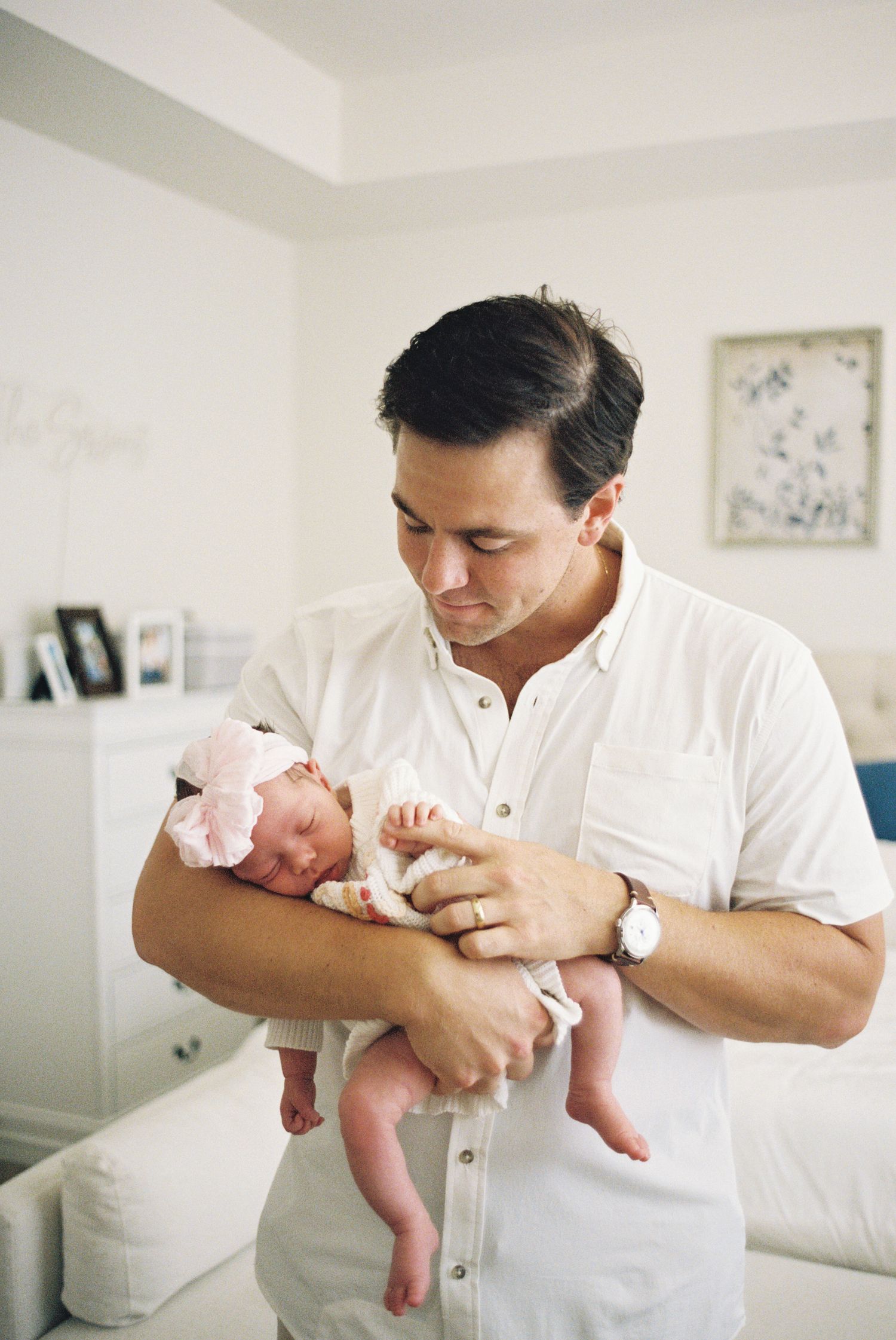 A father in a white shirt tenderly holds his newborn baby girl wearing a pink bow headband at home.