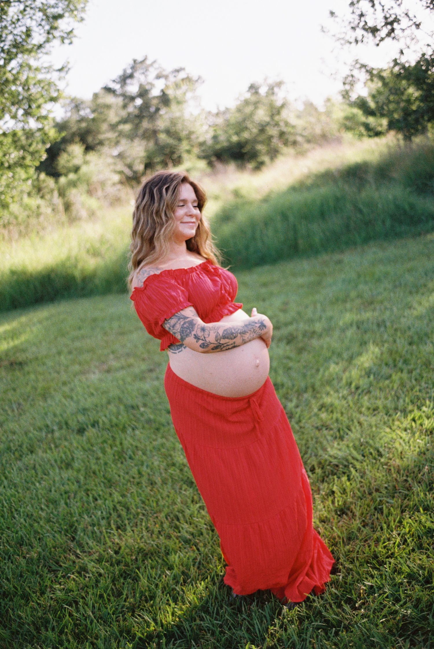 Pregnant woman in red off-shoulder maternity gown with tattoos poses outdoors on green grass field.