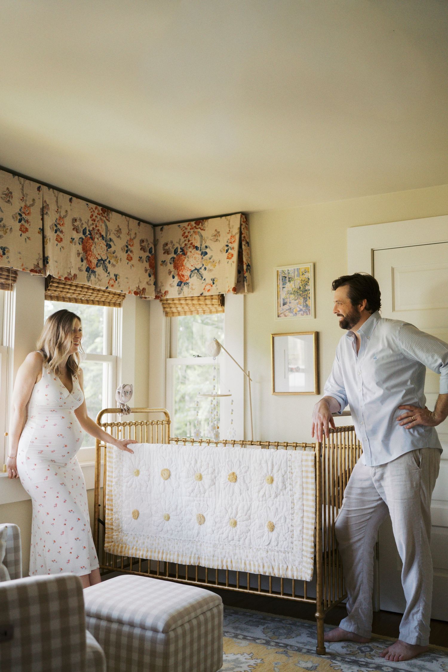 Pregnant woman and man standing by a gold crib in a cozy nursery with floral valances and yellow walls.