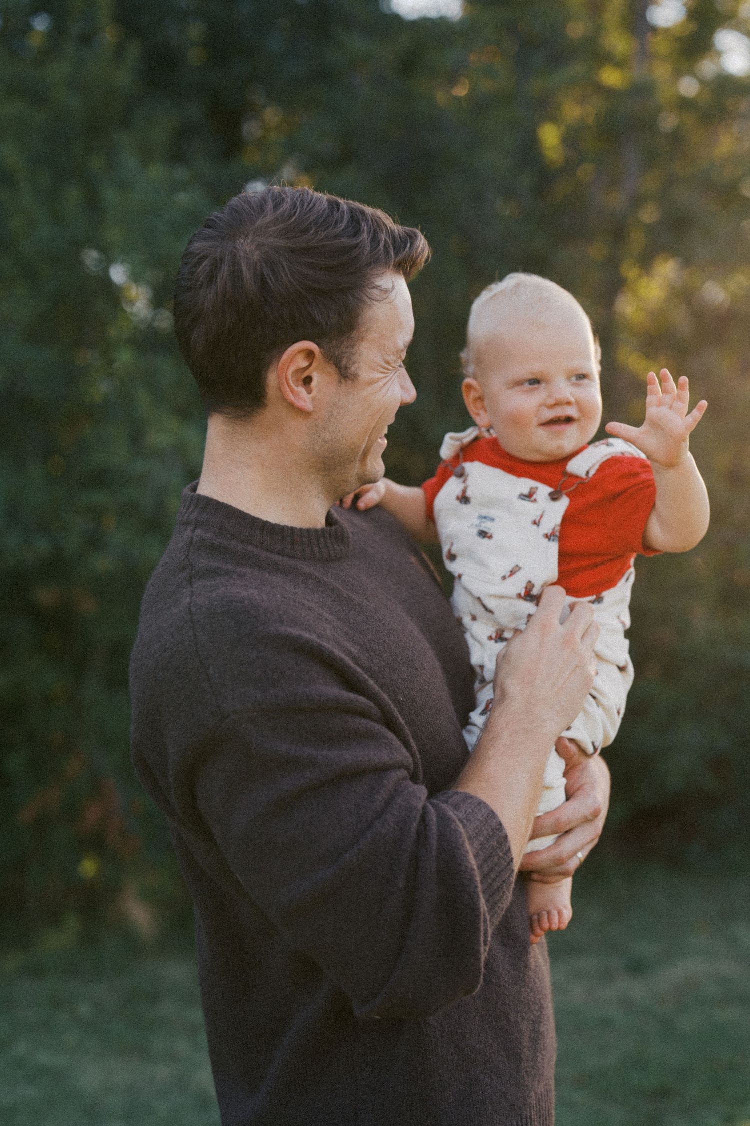 A tender moment between parent and infant in a brown sweater against a blurred outdoor background at sunset.