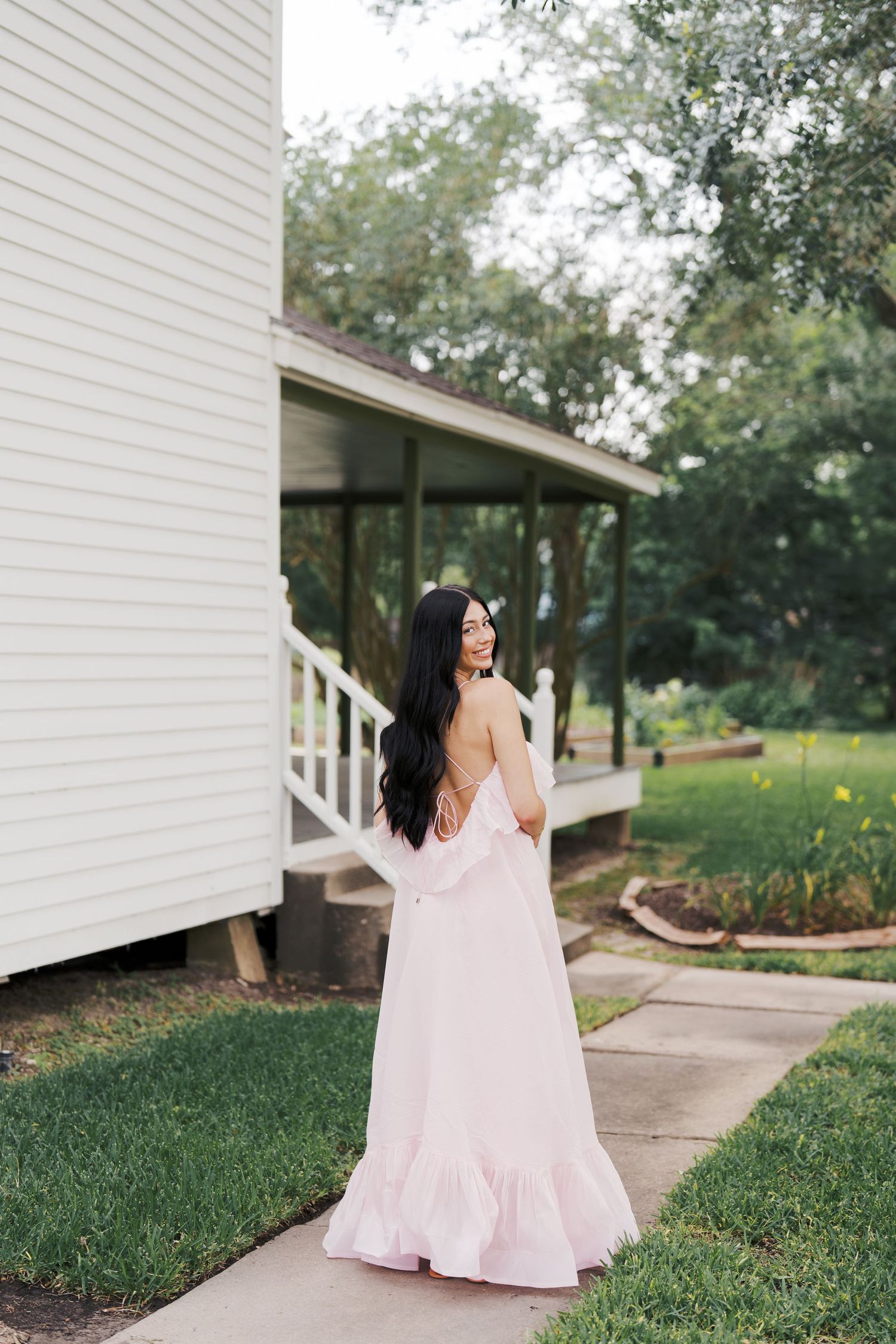 A woman in a pink maxi dress stands on a porch pathway.