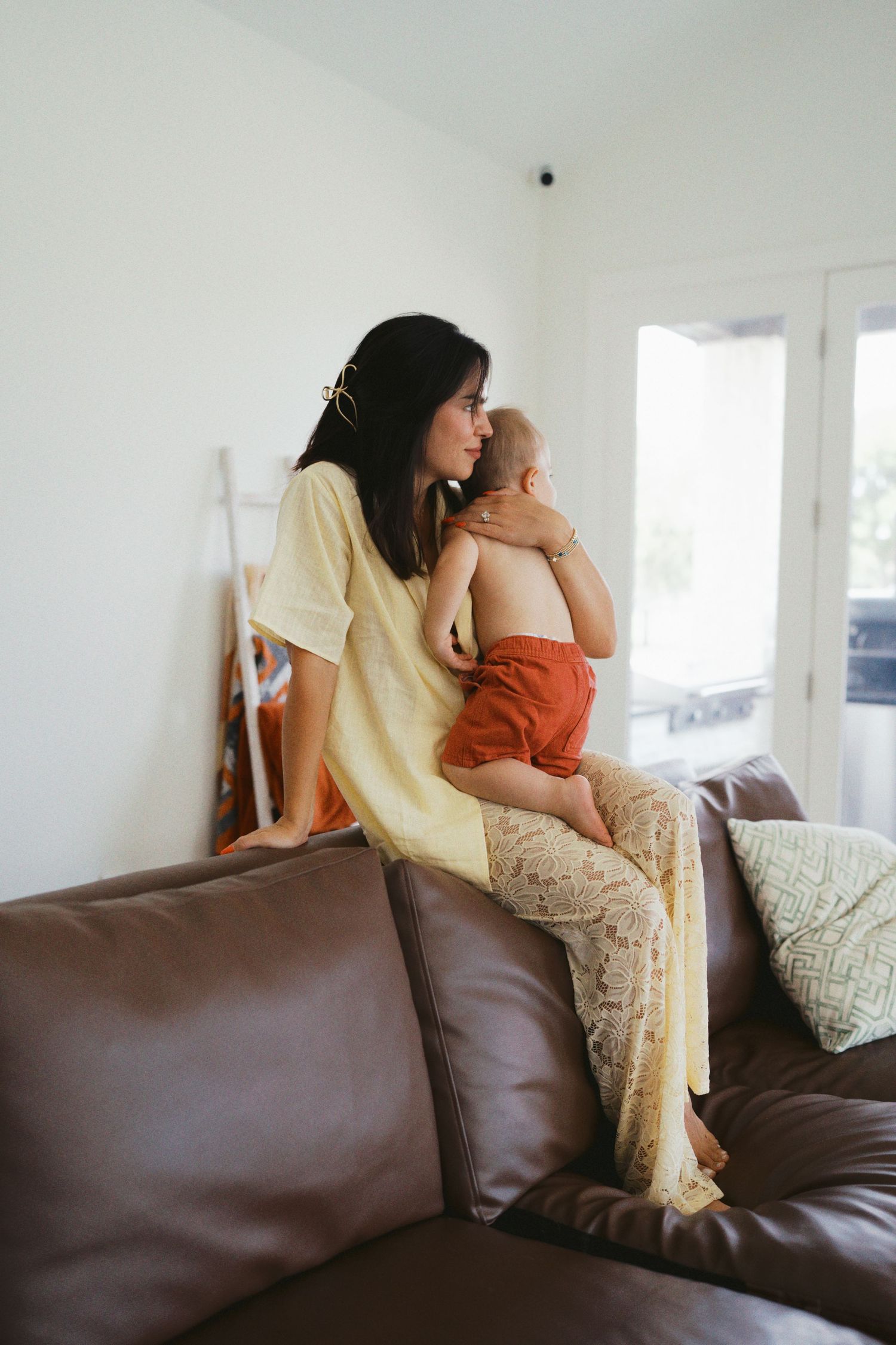 Young mother in yellow dress holding baby on brown leather sofa in bright living room.