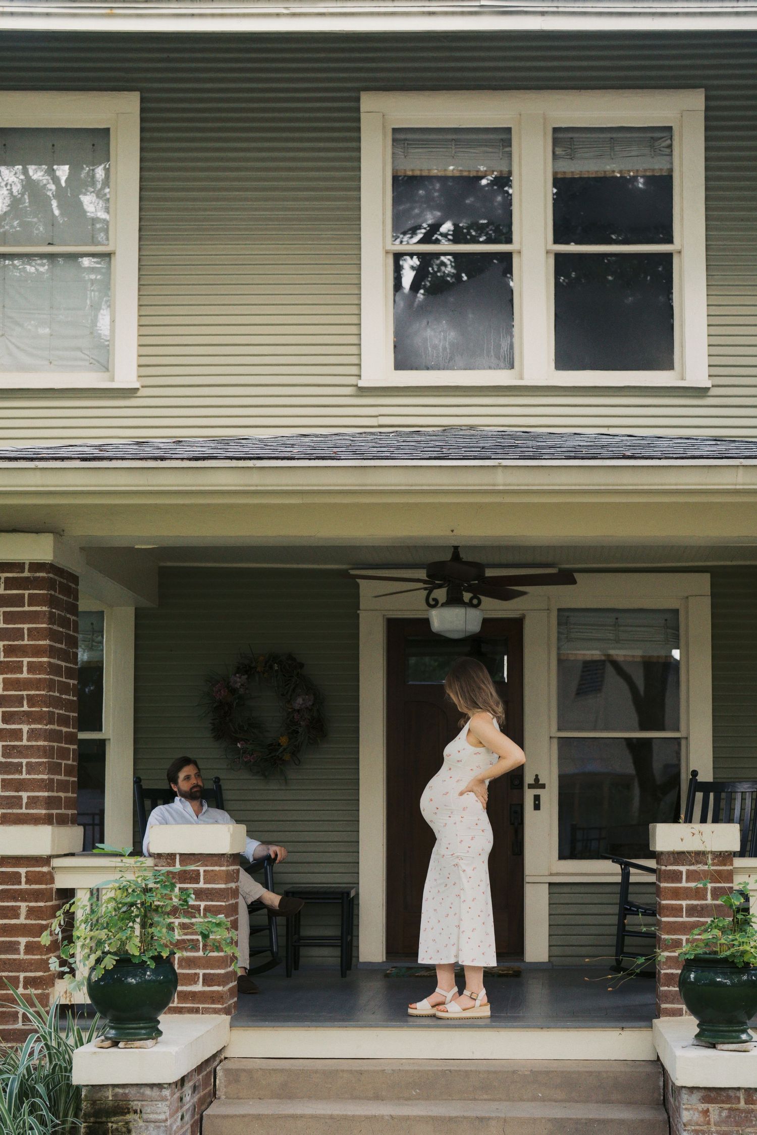 Pregnant woman in white dress stands on front porch of green house while man sits nearby.
