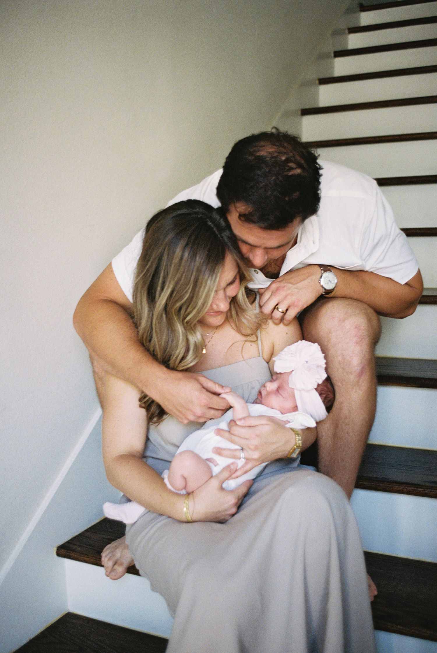 New parents sitting on staircase steps, lovingly gazing down at their newborn baby wrapped in white.
