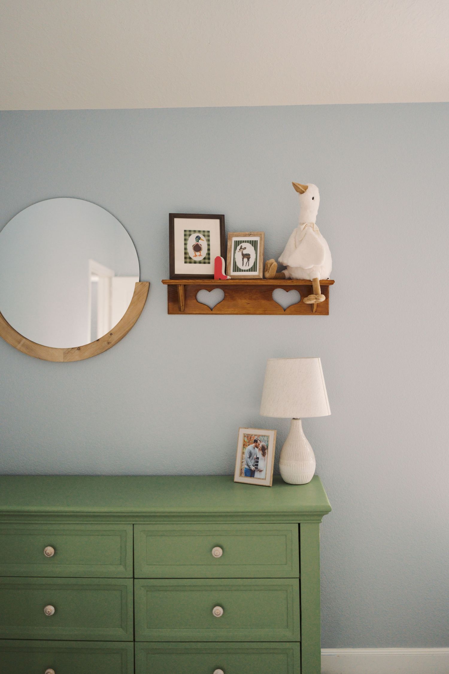 The image shows a green dresser with decorative items on top, including a round mirror, framed photos, and a decorative figurine.