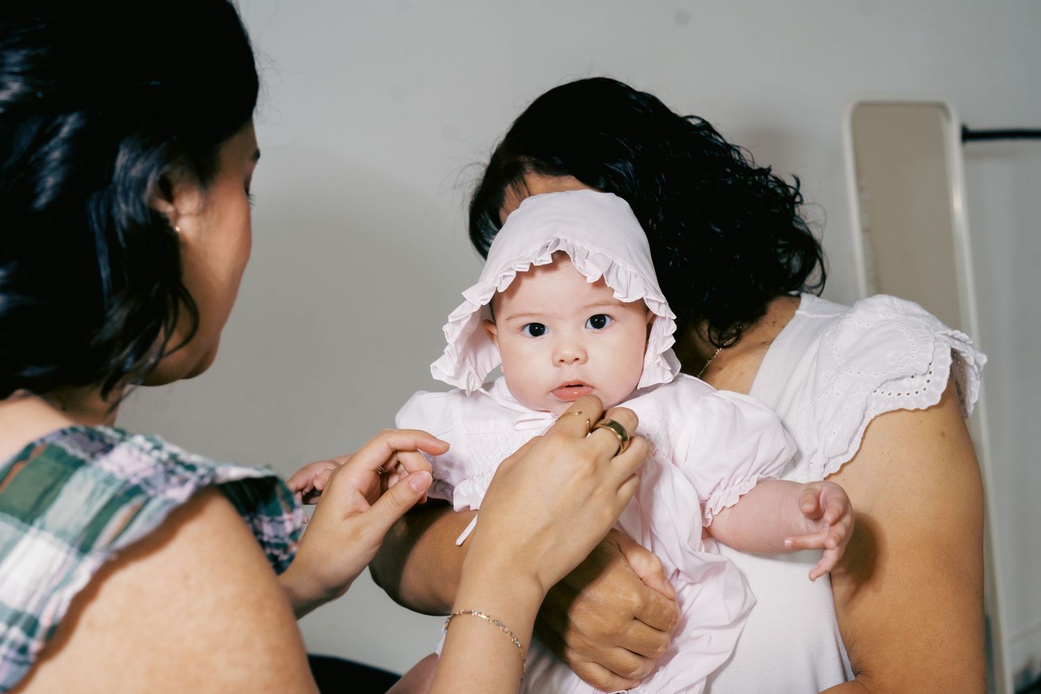 Two women dressing a cute baby girl in a pink bonnet and outfit, sharing a tender family moment.