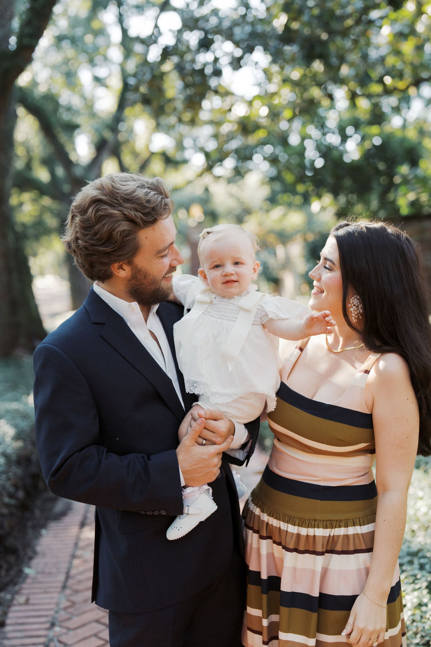 A family shares tender moments together in an outdoor setting with trees and brick pathways visible in the background.