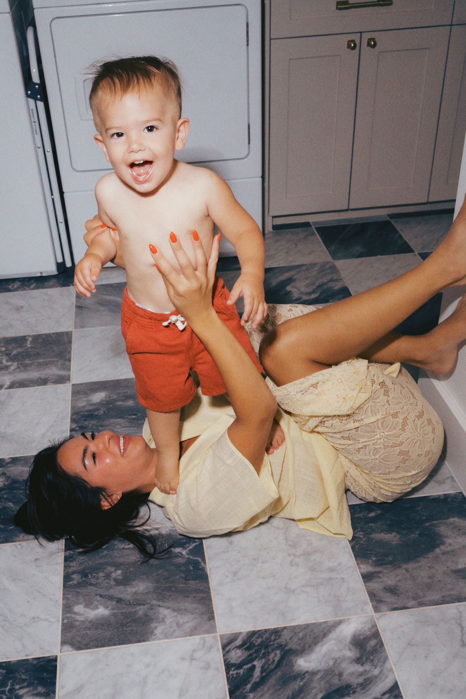 A mother lying on a checkered floor playfully holding her laughing toddler boy in red shorts.