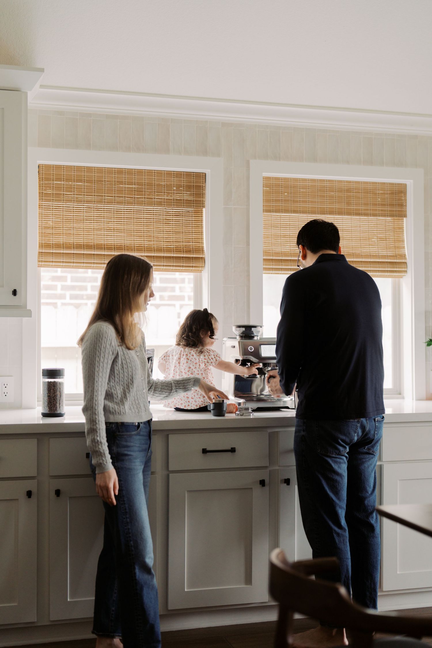 A family stands together in a bright kitchen with bamboo window shades and white cabinets while preparing food.