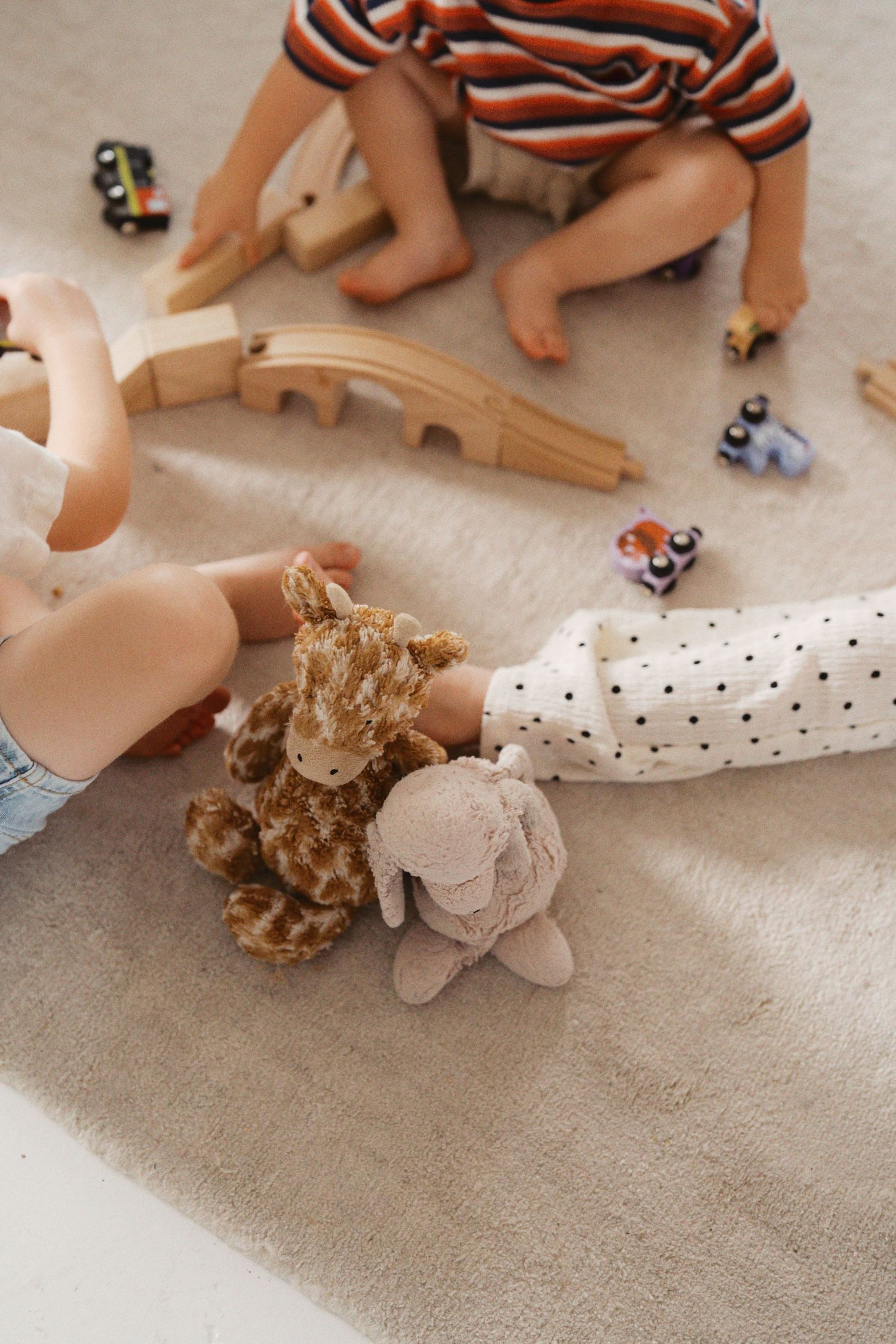 Two toddlers playing on a beige carpet with stuffed animals, wooden train set, and small toy cars.
