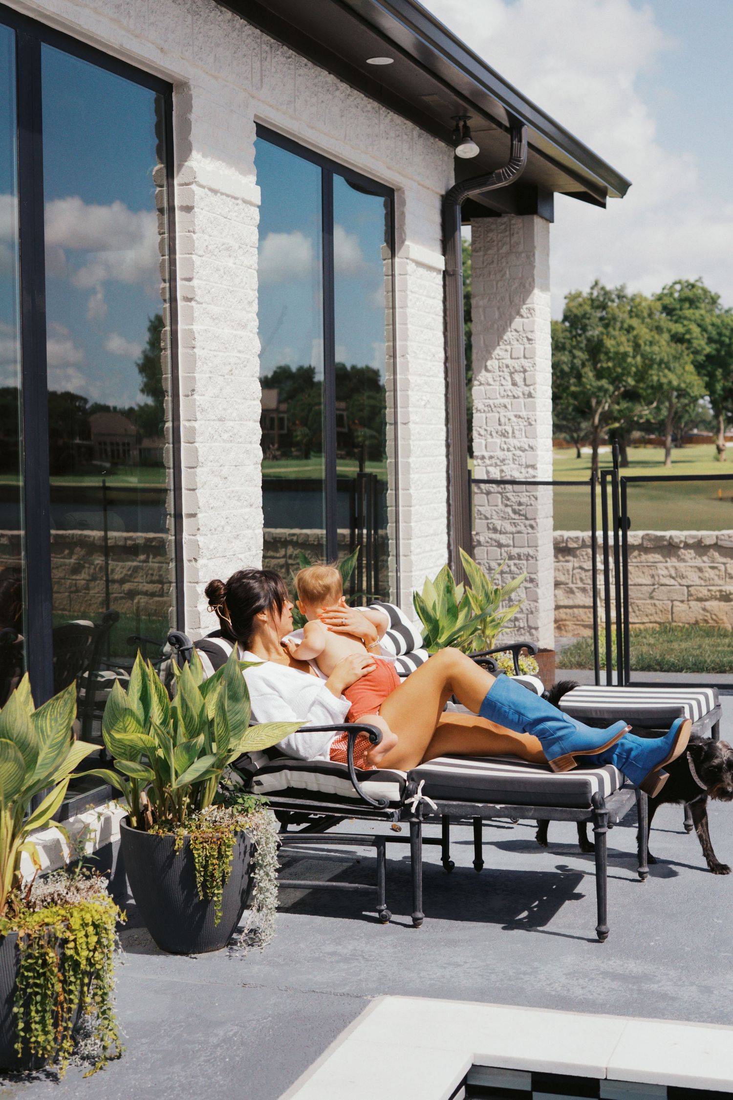 Woman relaxing on outdoor patio lounge chair holding a baby beside lush plants and modern home exterior.