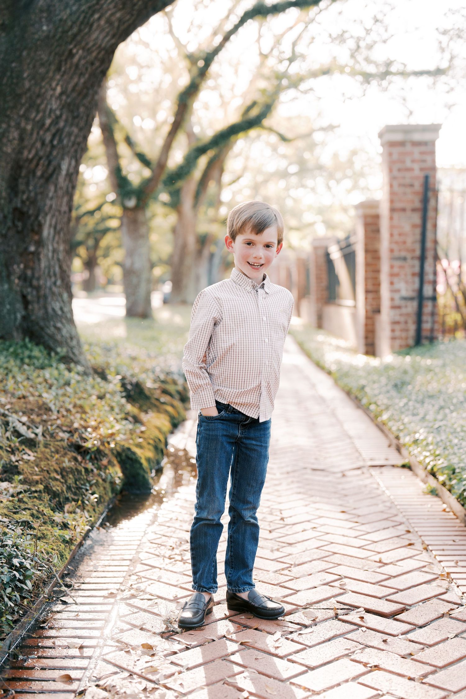 Young person standing on brick path under tree canopy wearing checkered shirt and jeans.
