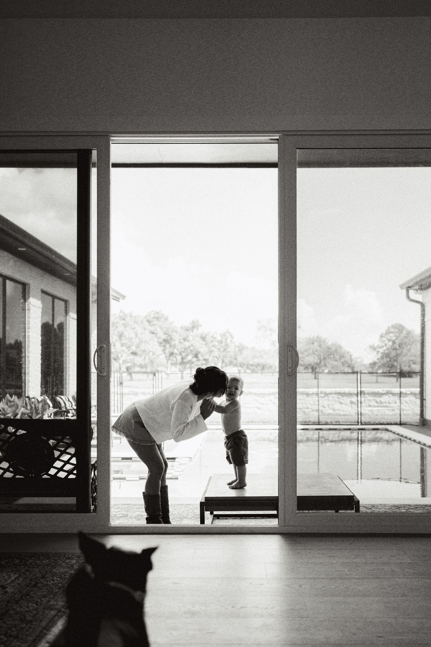 Black and white photo of parent bending down to embrace toddler in sliding glass doorway overlooking pool.