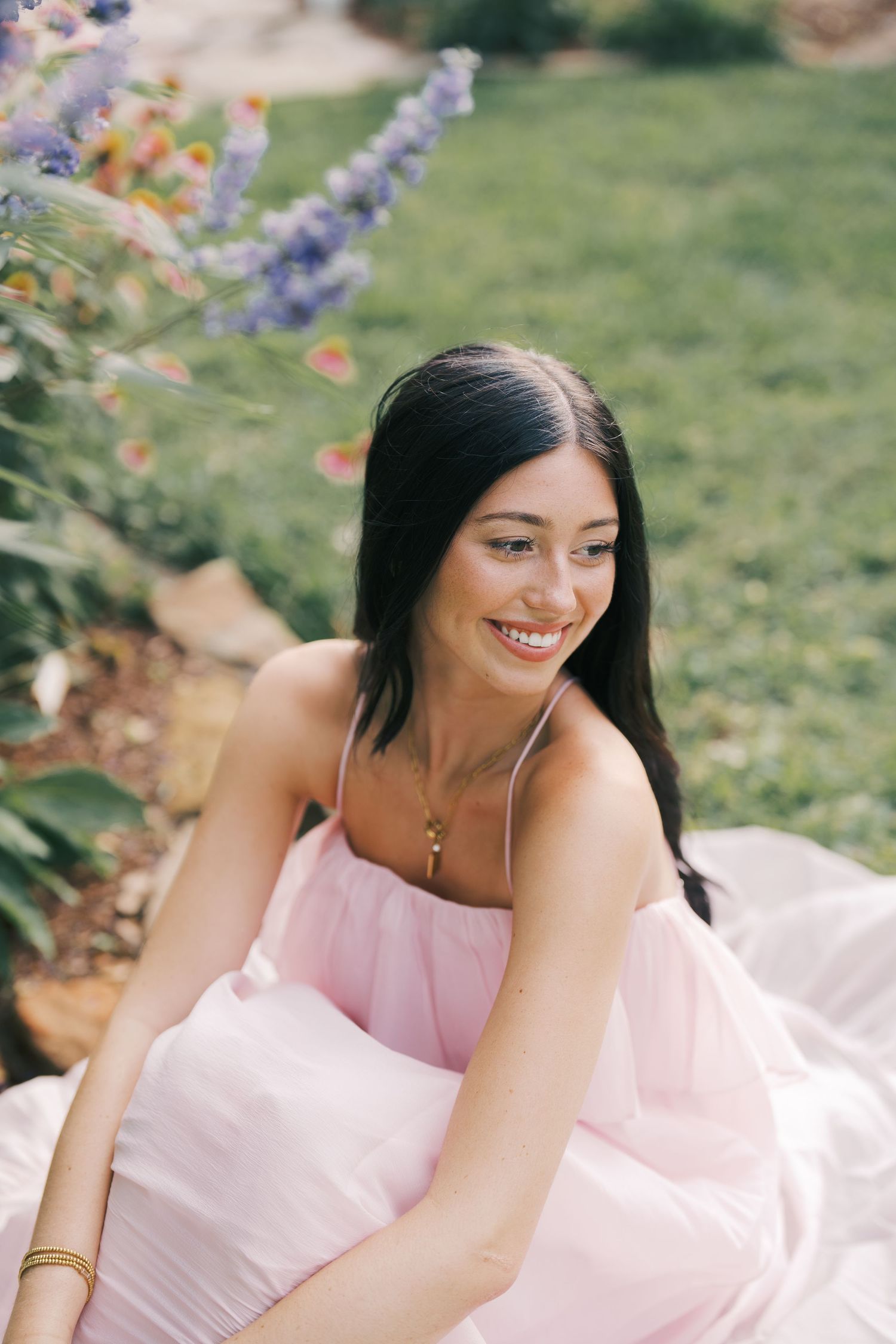 Portrait of person in pink dress sitting outdoors surrounded by colorful flowers.
