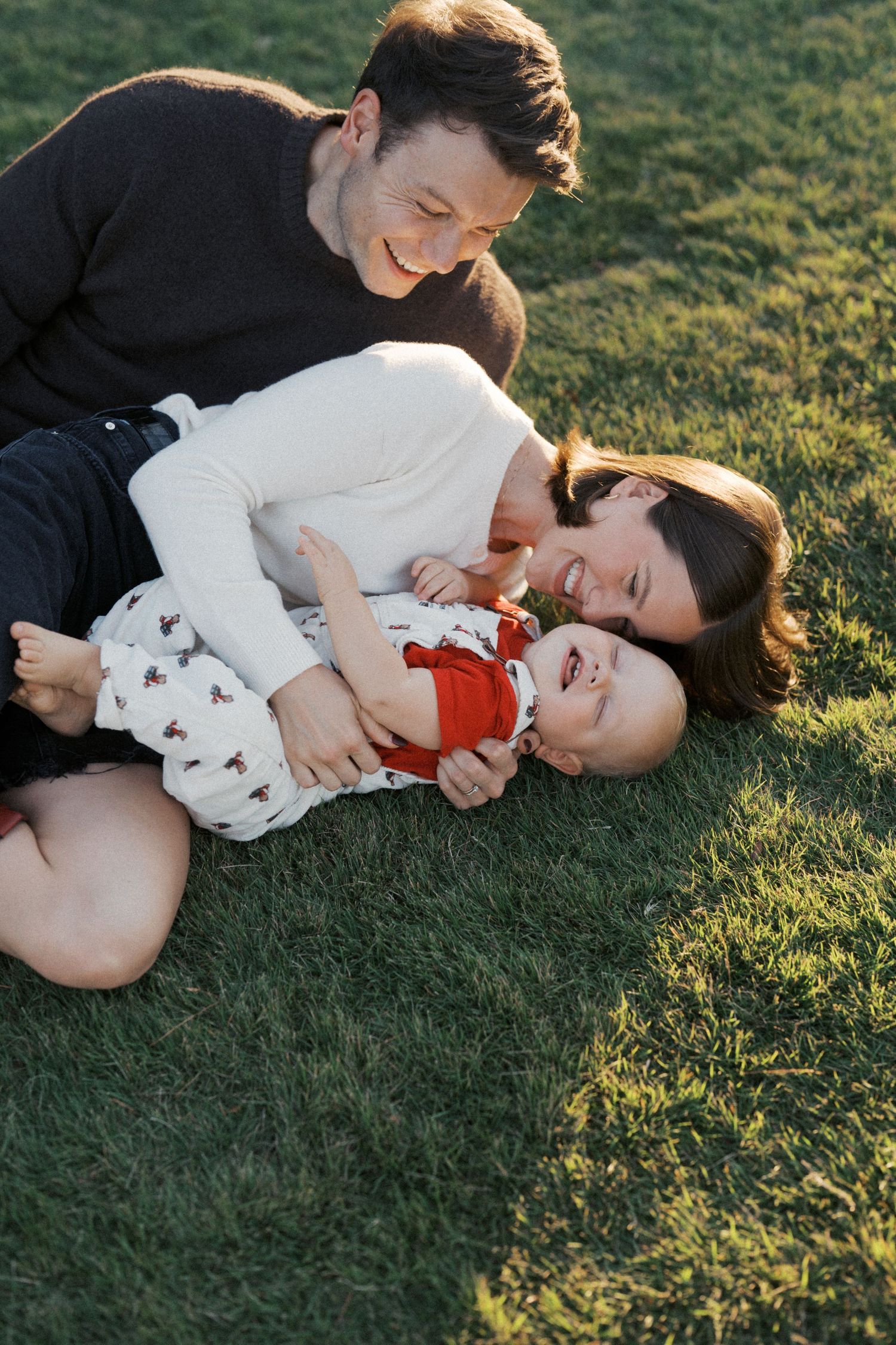 A playful couple lies together on green grass during golden hour sunlight.