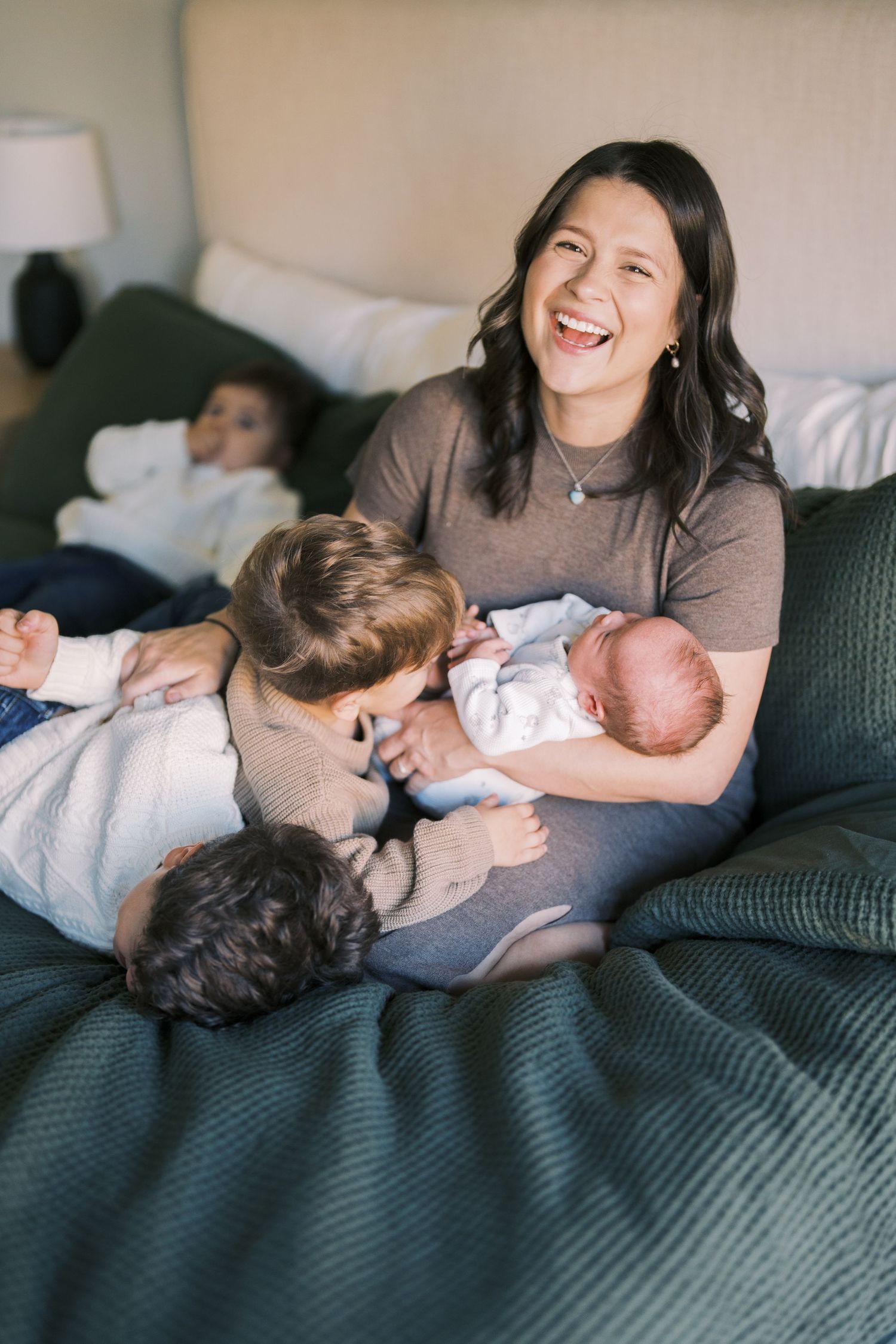 A joyful mother laughs while holding a newborn on a bed, surrounded by her young children in a cozy bedroom.