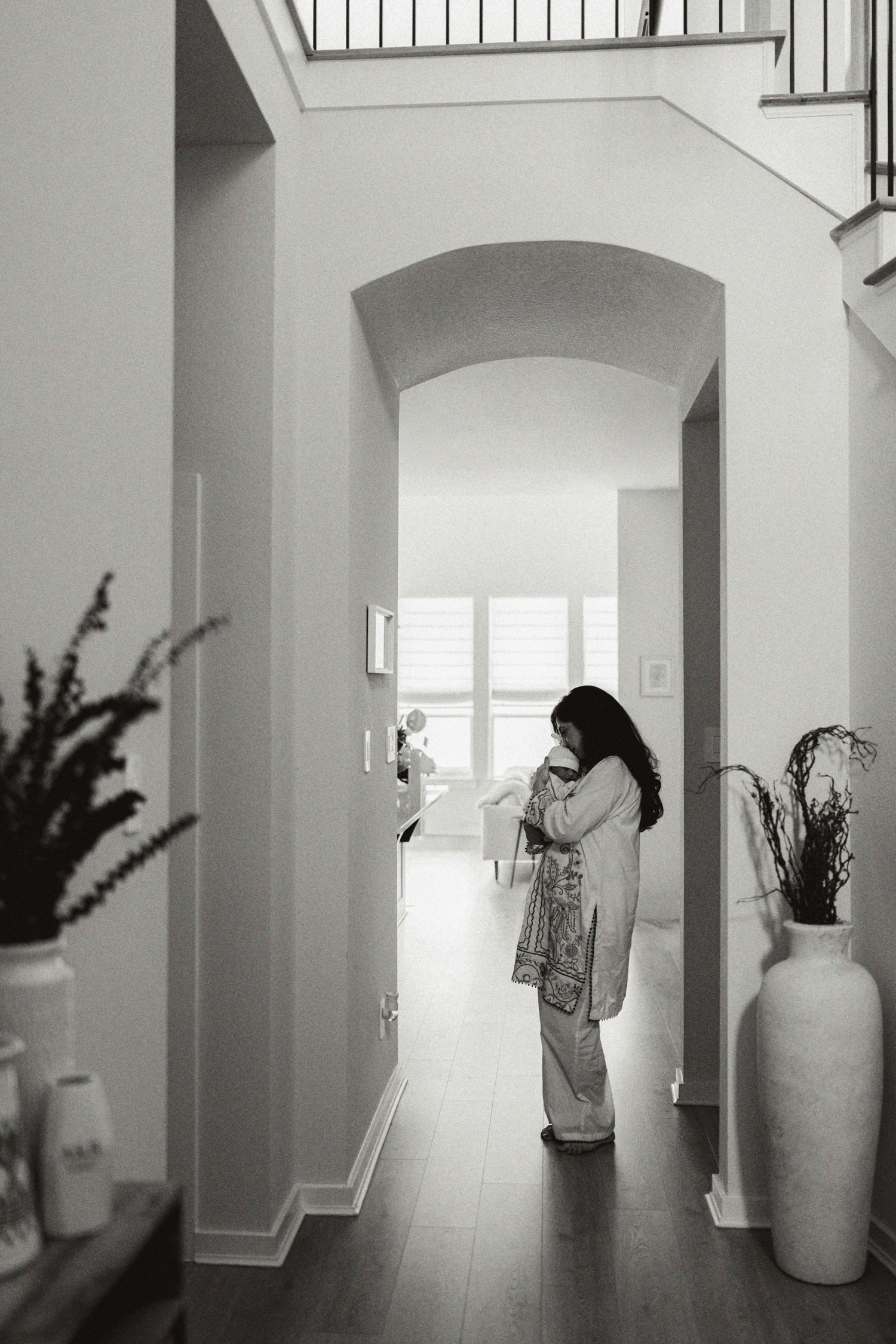 Black and white photo of a woman standing in a hallway with an arched doorway, holding a bag in a modern home.