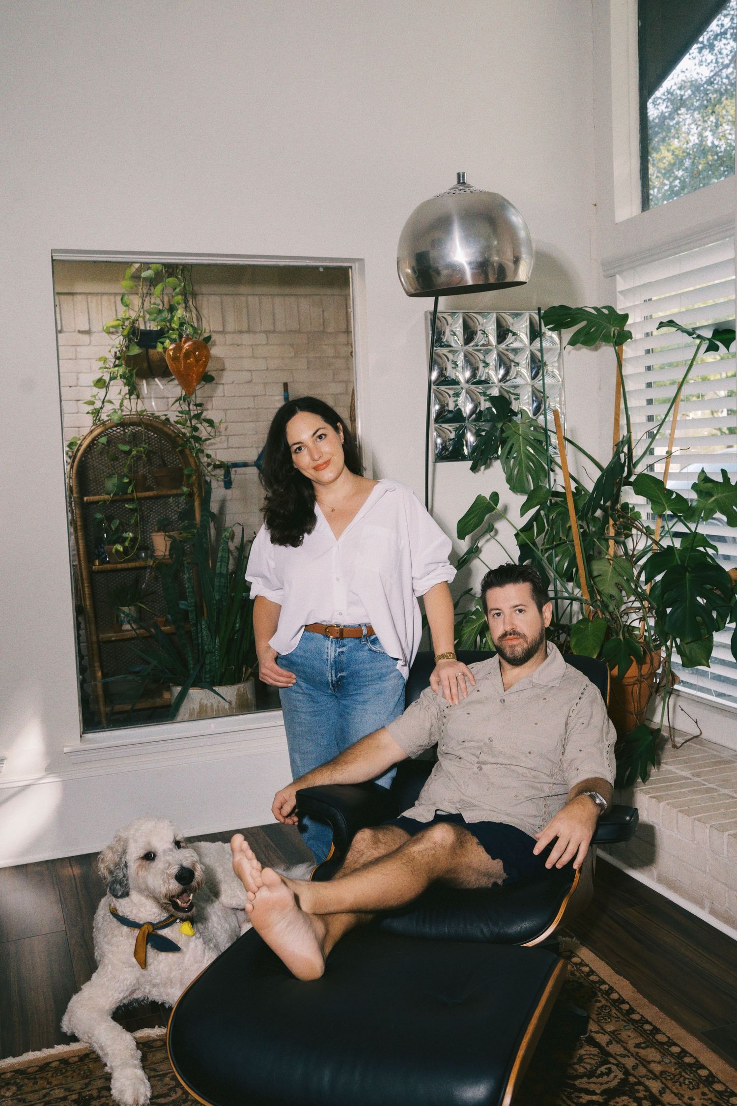 Couple posing with white fluffy dog in bright modern living room with plants and pendant light.