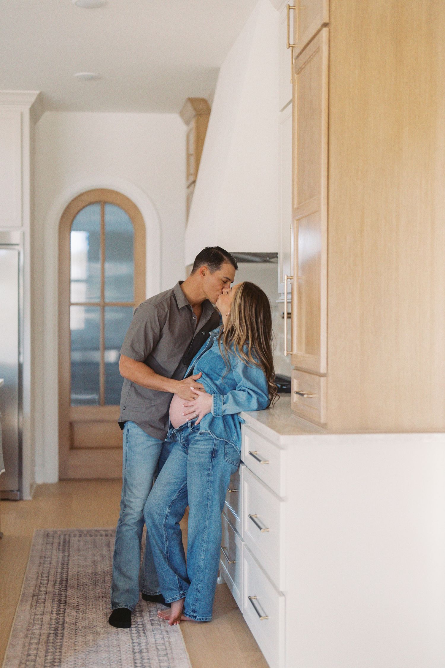 A couple shares an intimate moment while standing in a bright, modern hallway with white walls and light wood floors.
