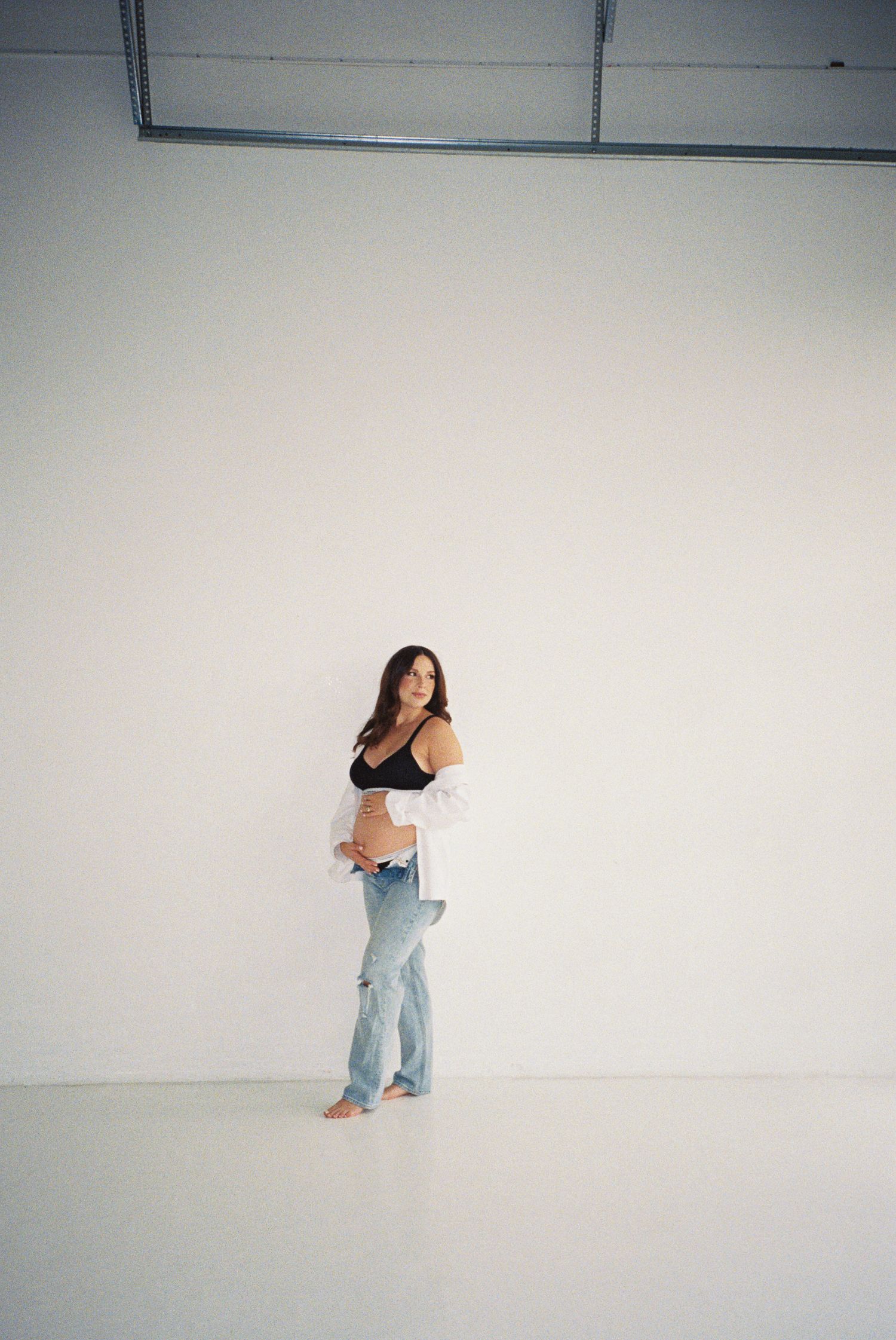 Pregnant woman in black crop top, white shirt, and jeans posing barefoot against a white studio wall.