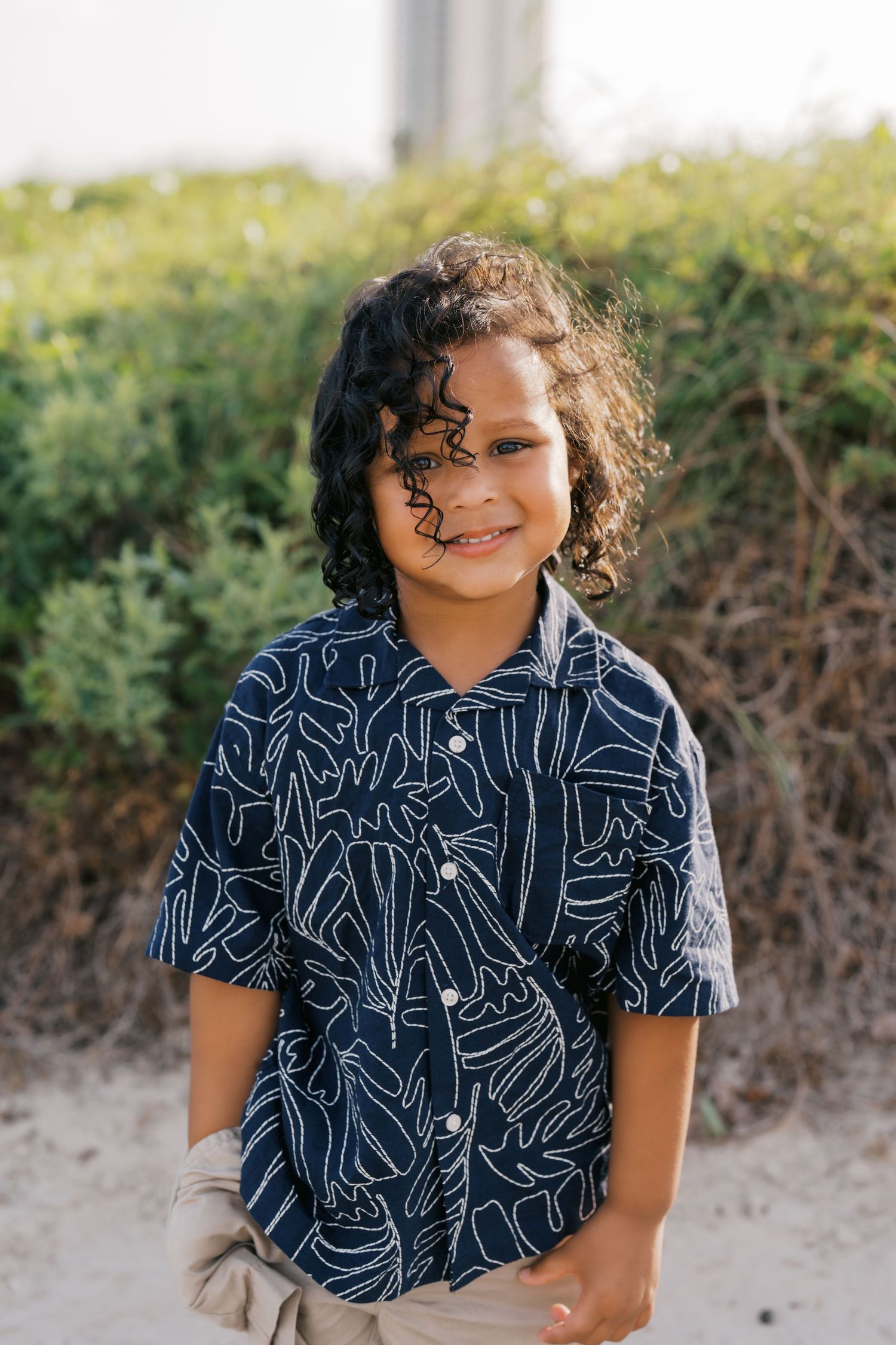 Smiling young boy with curly hair wearing a navy blue patterned short-sleeve shirt outdoors.