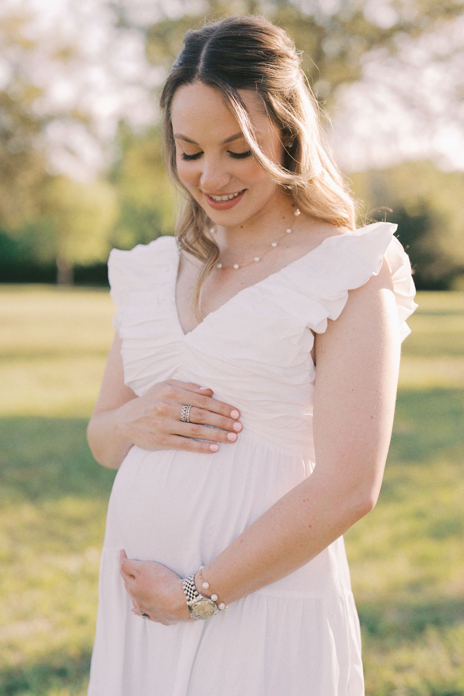 Expectant mother in flowing white dress smiles while cradling her baby bump in a sunlit outdoor setting.