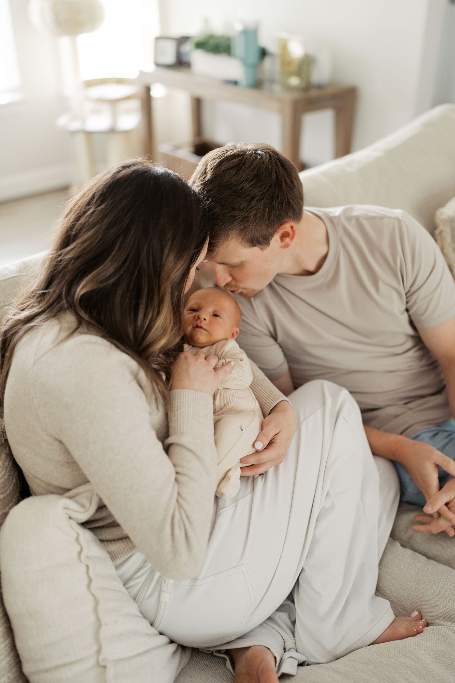 Parents tenderly kissing their newborn baby while sitting together on a cozy white sofa at home.