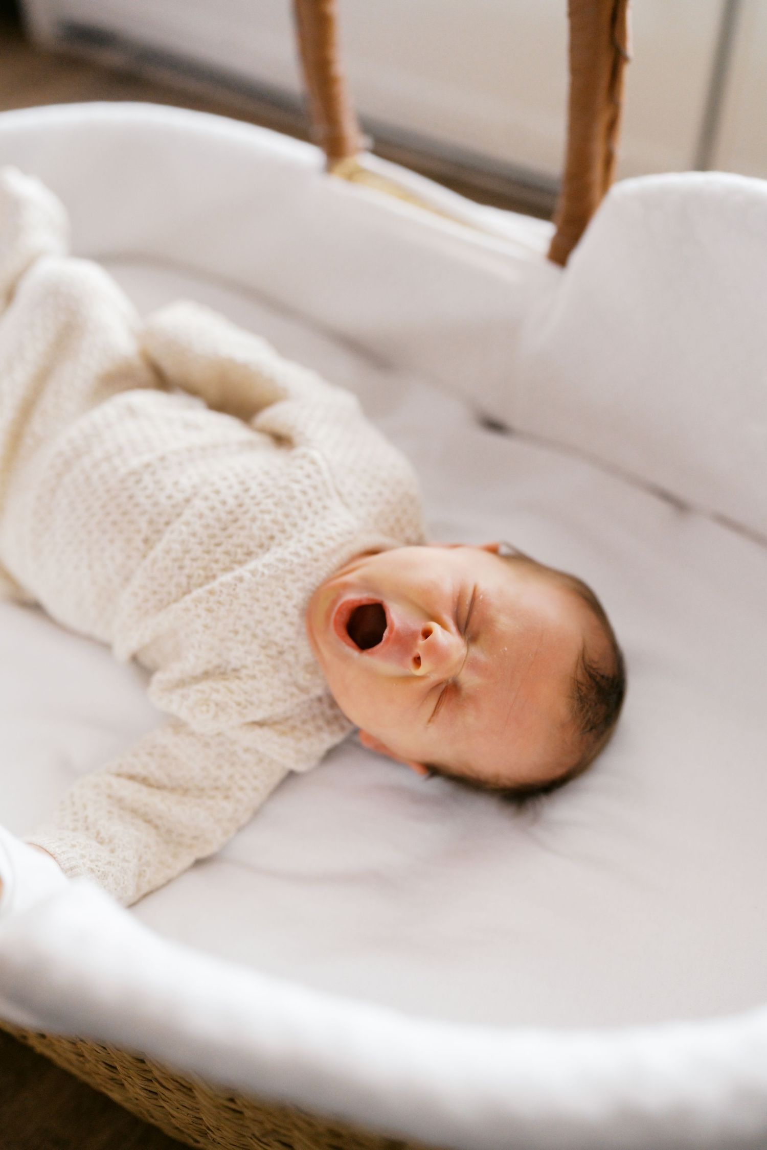 A newborn baby yawning while swaddled in a white knit blanket, lying in a wicker Moses basket.