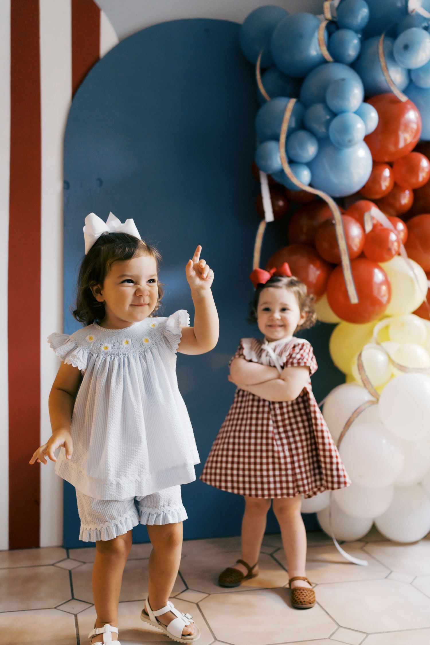 Two toddler girls in festive dresses pose near colorful balloons at a birthday party celebration.