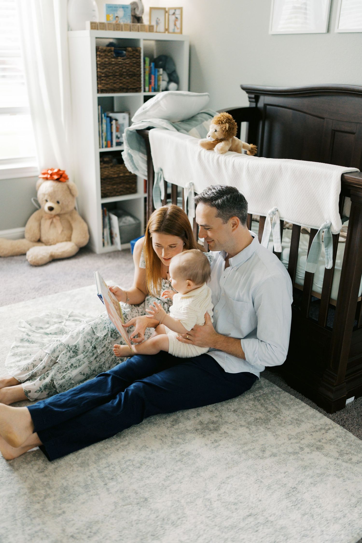 Young family sitting on nursery floor reading to baby near dark wood crib with teddy bears in background.