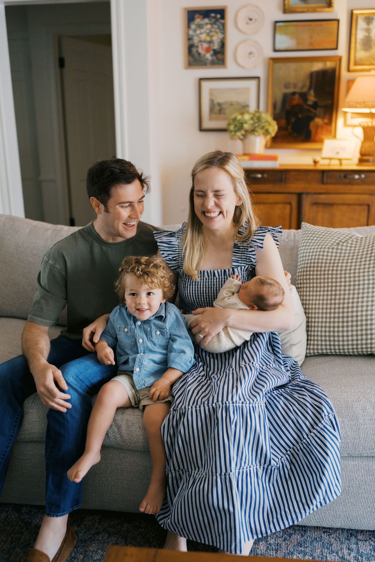 Happy family of four sitting on a cozy sofa, parents smiling with toddler and newborn baby in a warm home.