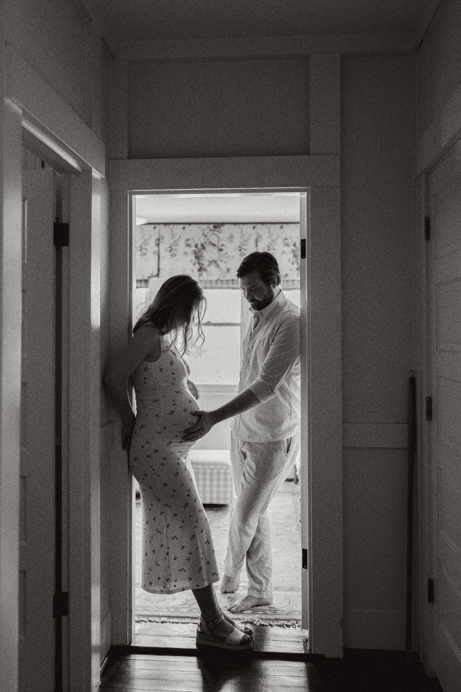 Black and white maternity photo of couple in doorway, man tenderly touching pregnant woman's belly in soft natural light.