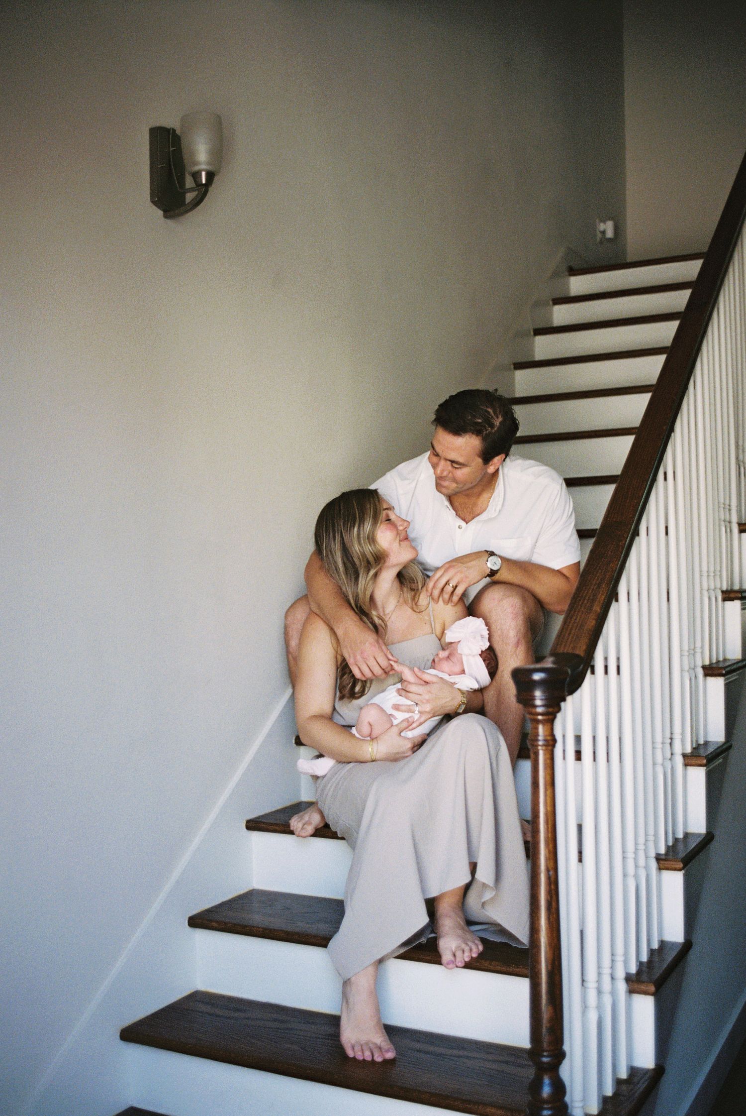 A loving couple sits on wooden stairs, cradling their newborn baby together in a warm home setting.