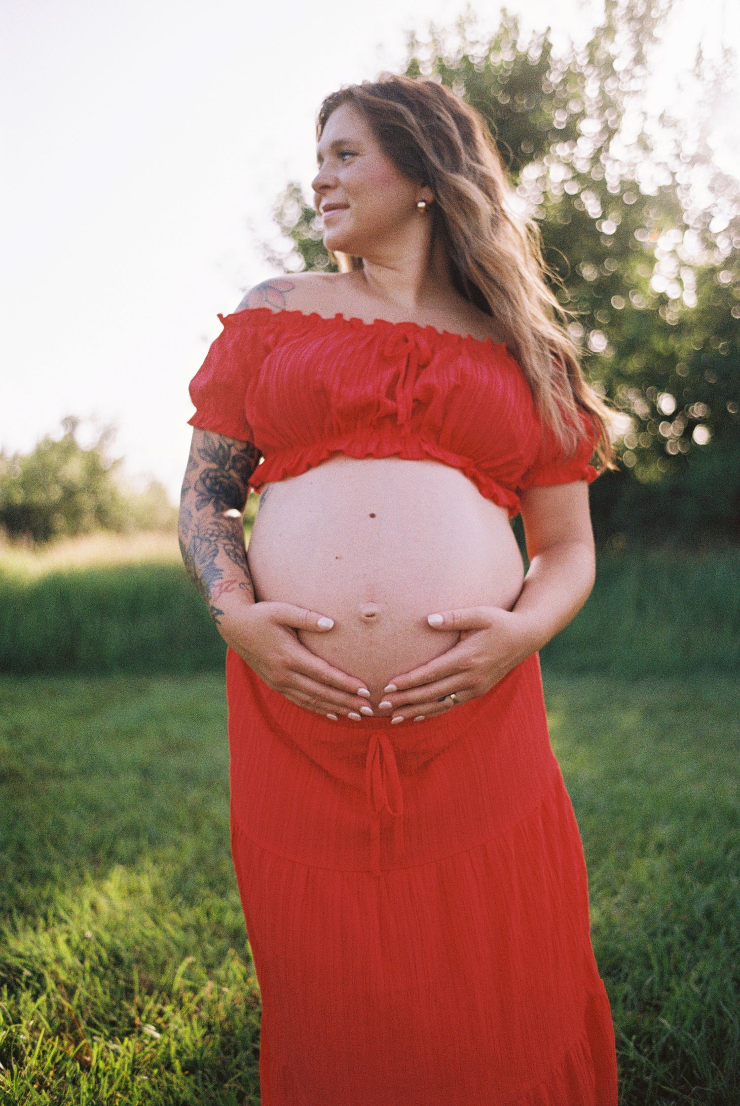 Pregnant woman in red off-shoulder dress holding baby bump outdoors in green field during golden hour.