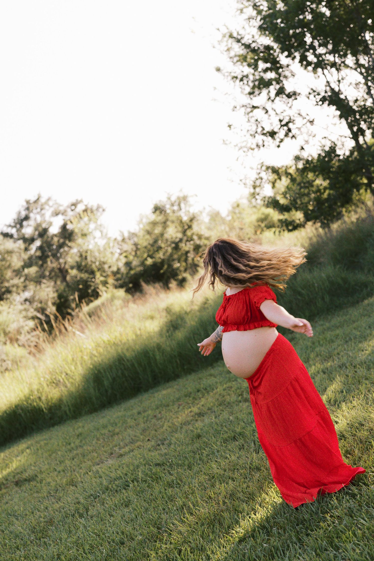 Pregnant woman in red off-shoulder dress twirling on green grass with trees in background during golden hour.