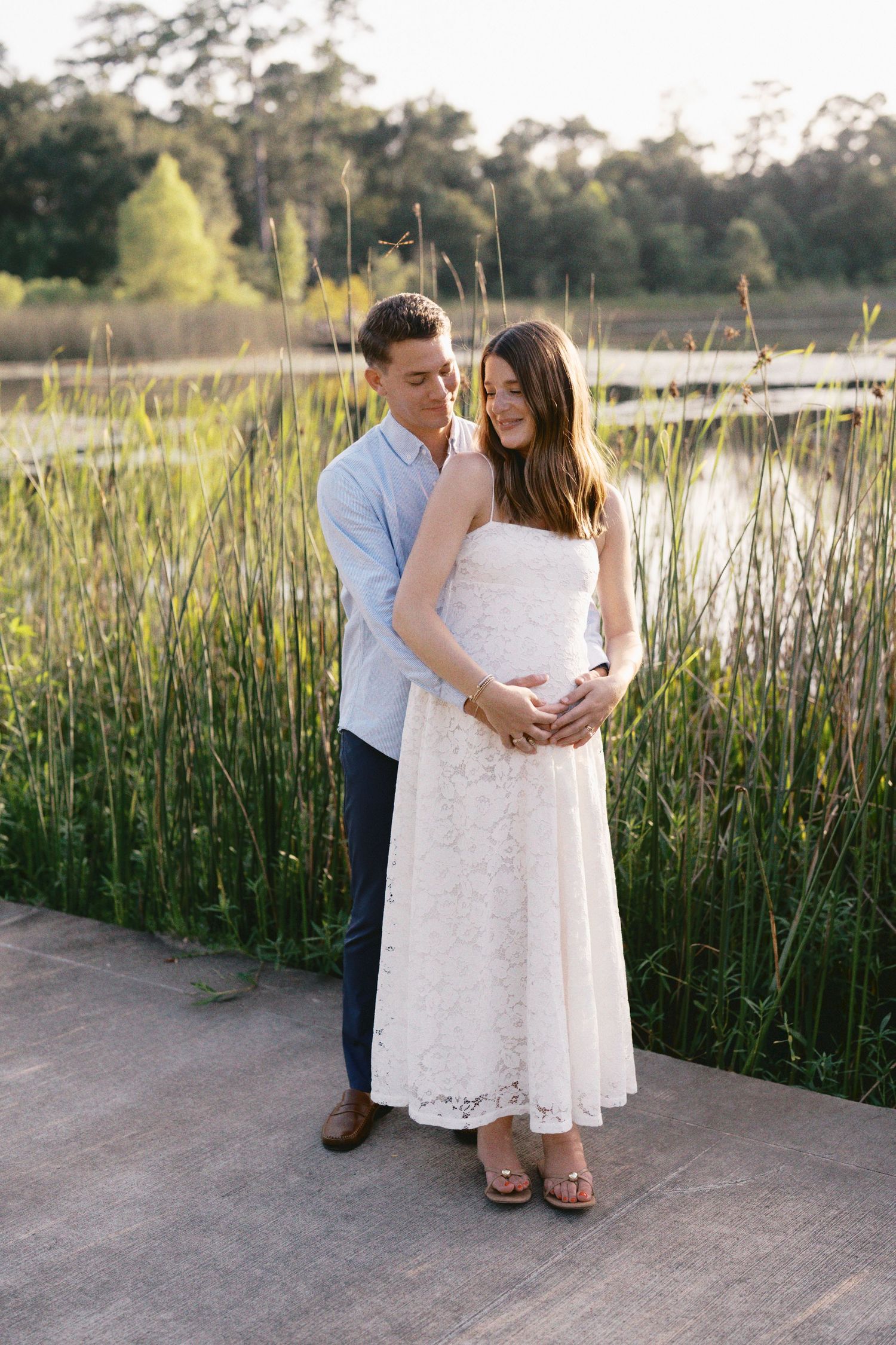 Couple embracing outdoors near a pond, woman in white lace dress with hands on baby bump, golden hour light.
