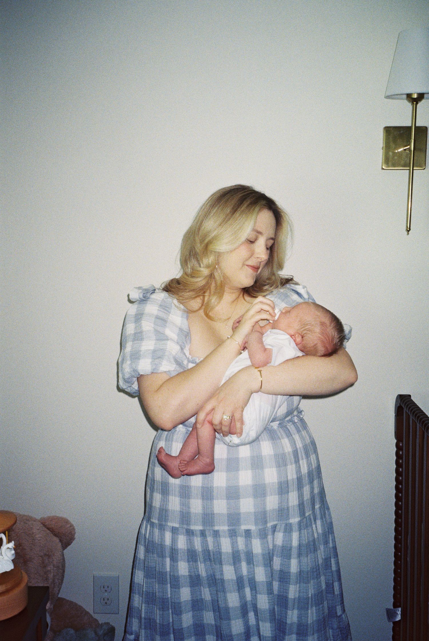 A blonde woman in a blue gingham dress tenderly holds a newborn baby in a warmly lit room.