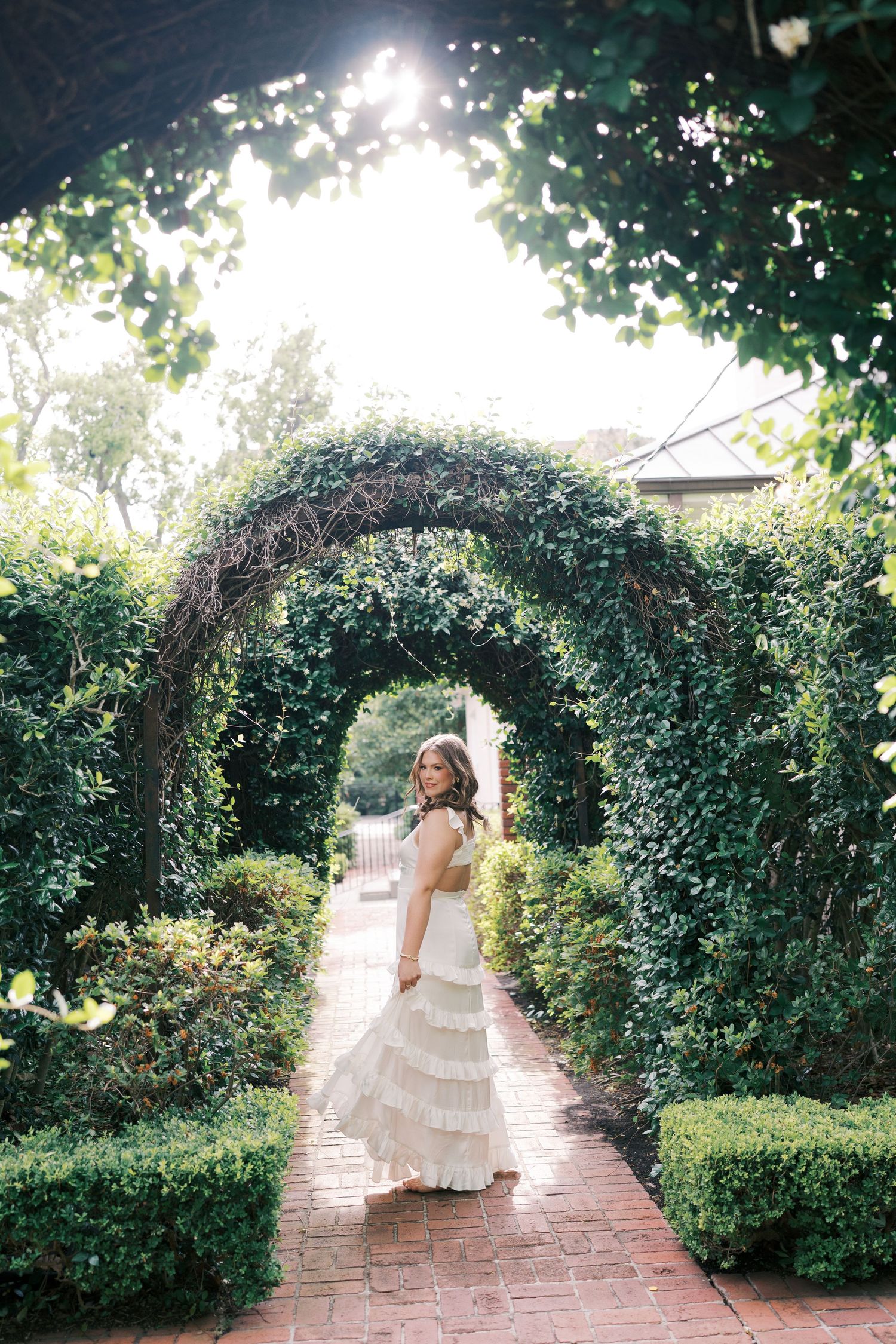Bride in tiered wedding dress poses under vine-covered garden archway with sunlight peeking through.