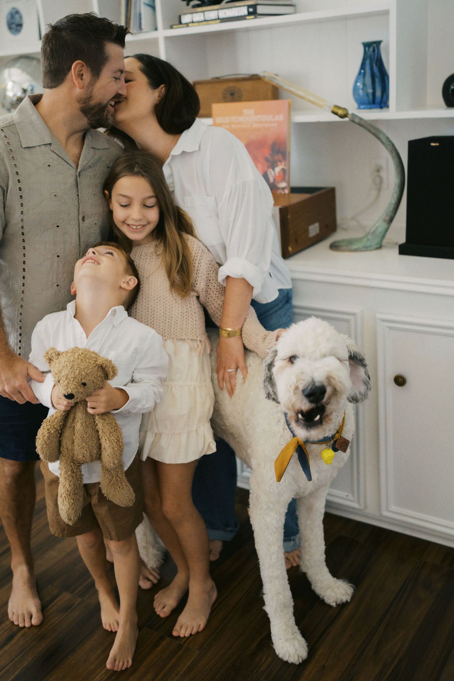 Happy family with two kids and a white fluffy dog posing together indoors, parents kissing while children smile and hug.