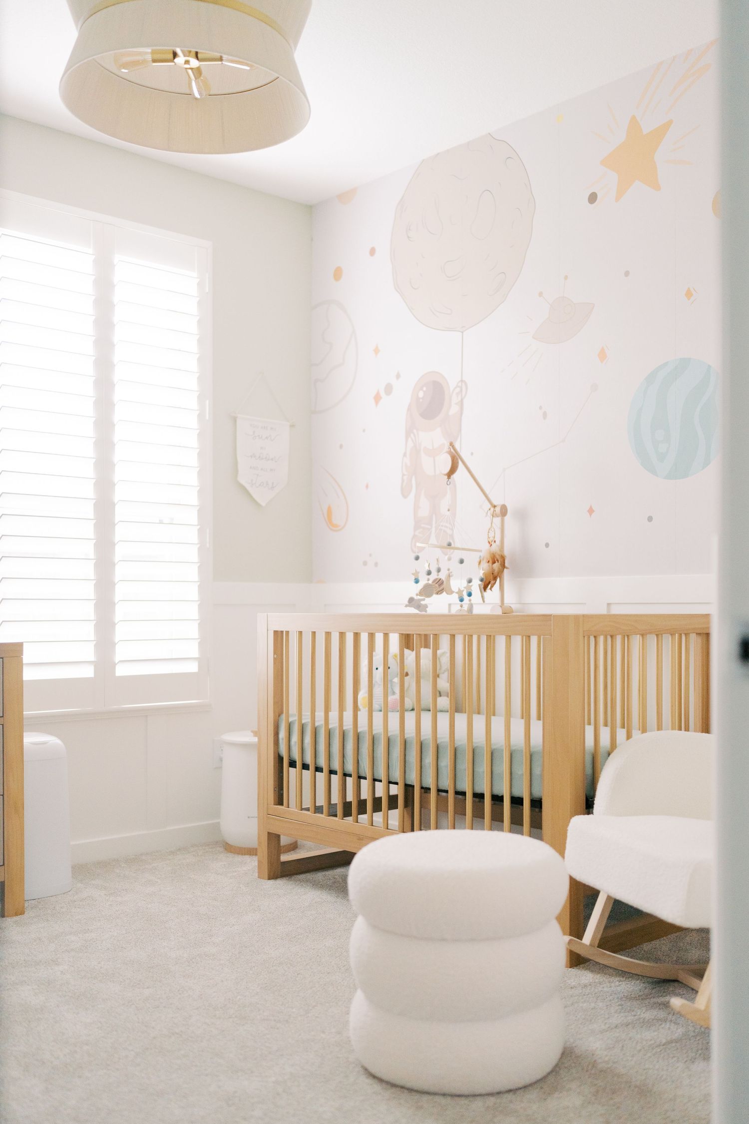 Modern nursery with neutral decor featuring a wooden crib, white walls adorned with celestial designs, and minimalist furniture.