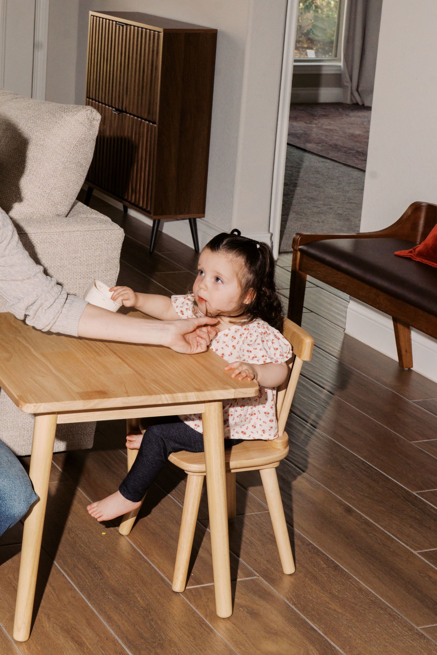 A young girl sitting at a small wooden table in a cozy living room with a wooden cabinet and leather sofa in the background.