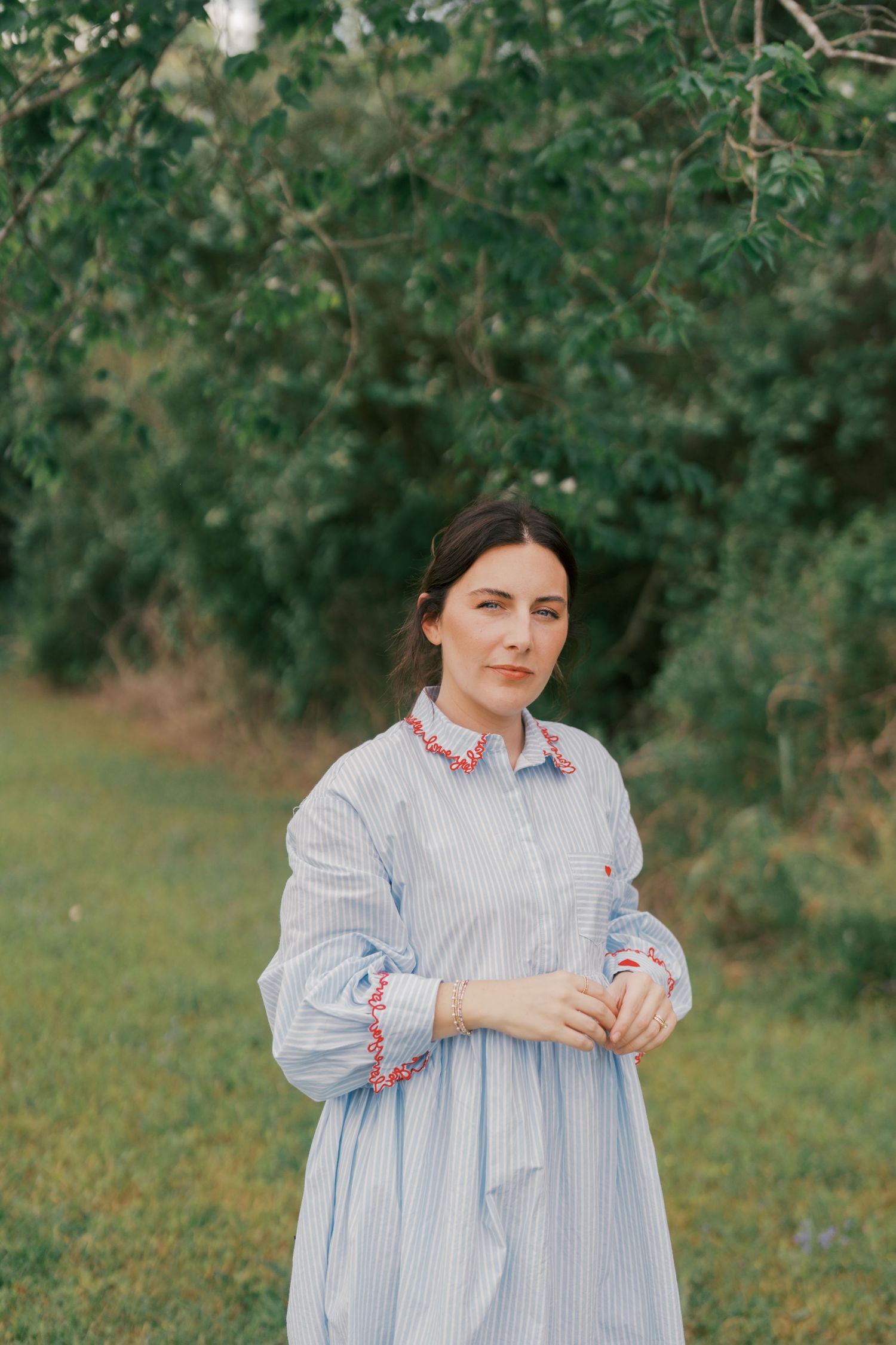 Woman in blue striped dress with red embroidered collar stands outdoors among green trees.