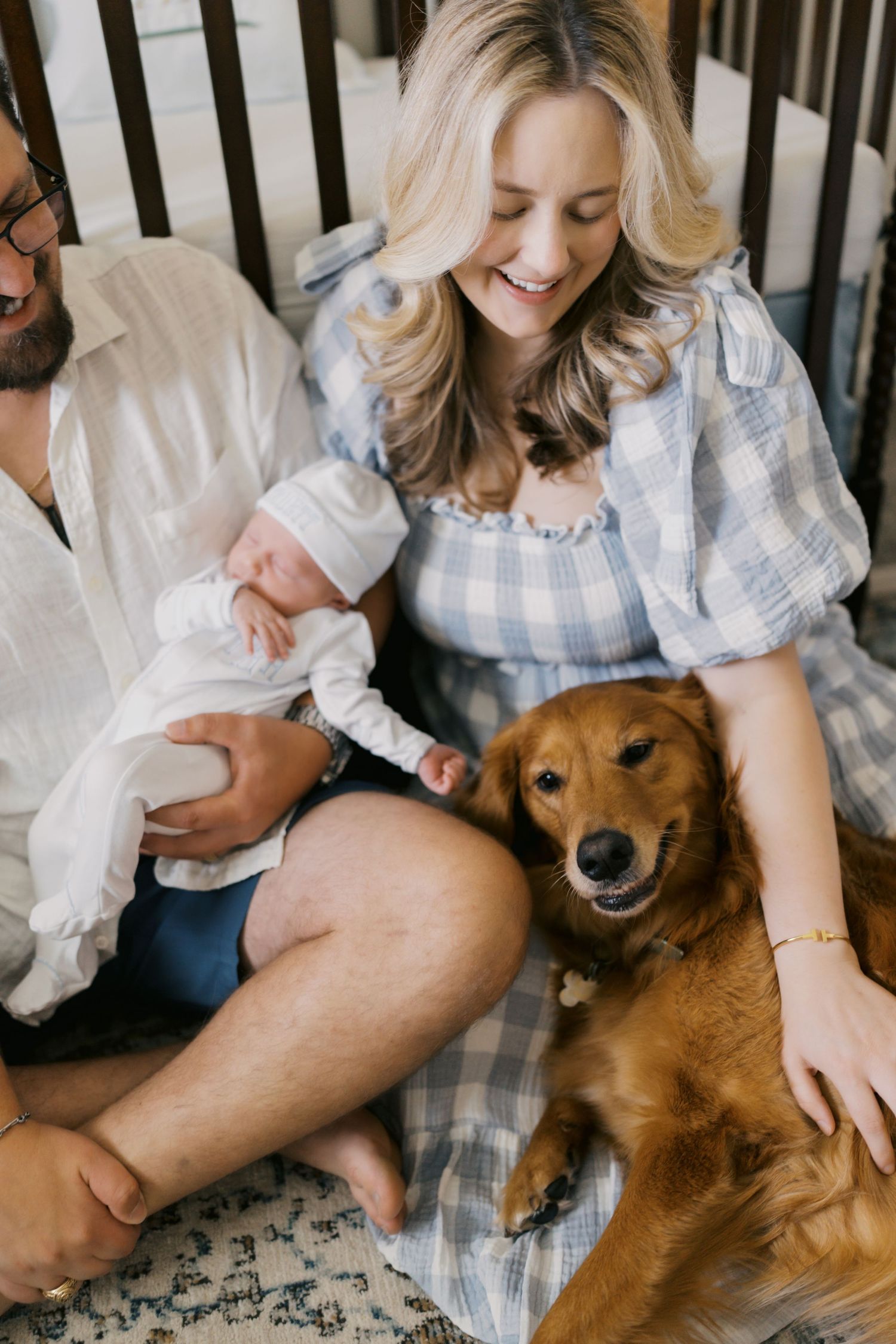 A smiling couple sits with their newborn baby and golden retriever dog in a cozy nursery setting.